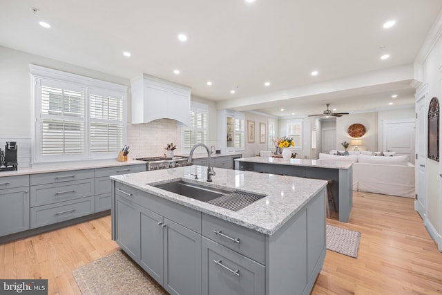 kitchen featuring sink, a kitchen island with sink, light stone countertops, custom range hood, and light hardwood / wood-style flooring