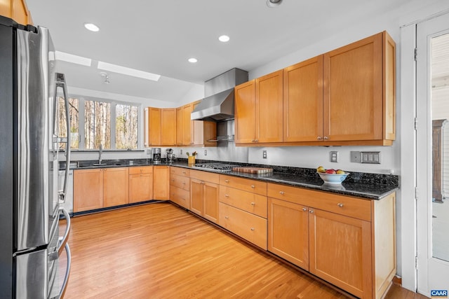 kitchen featuring appliances with stainless steel finishes, sink, dark stone countertops, wall chimney range hood, and light hardwood / wood-style flooring