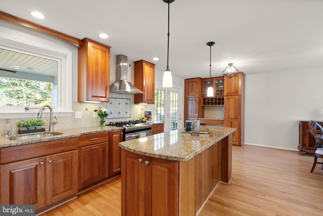 kitchen with a sink, stainless steel range with gas cooktop, wall chimney exhaust hood, brown cabinetry, and a healthy amount of sunlight