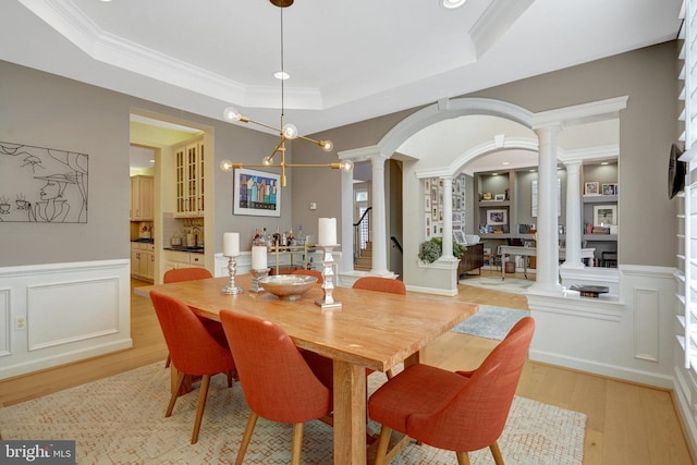 dining room with light hardwood / wood-style flooring, built in shelves, a raised ceiling, crown molding, and ornate columns