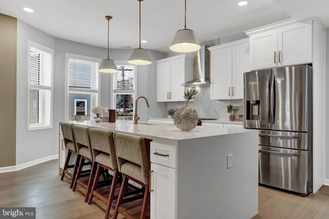 kitchen featuring wall chimney range hood, sink, a kitchen island with sink, white cabinetry, and stainless steel refrigerator with ice dispenser