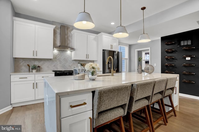 kitchen with stainless steel refrigerator with ice dispenser, a kitchen island with sink, white cabinets, and wall chimney exhaust hood