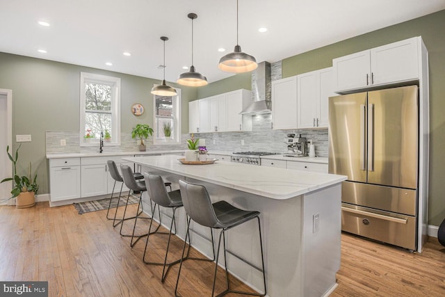 kitchen with white cabinetry, appliances with stainless steel finishes, wall chimney range hood, light wood-type flooring, and a kitchen bar