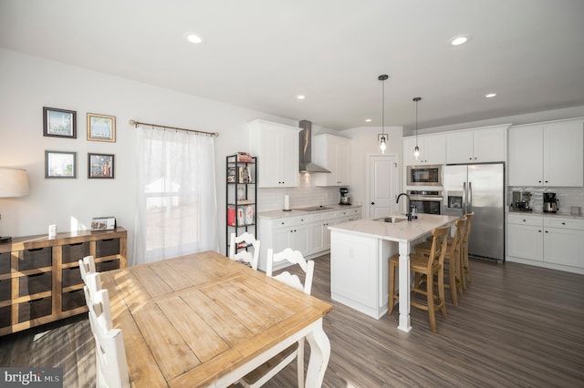 kitchen with sink, stainless steel appliances, an island with sink, decorative light fixtures, and wall chimney exhaust hood