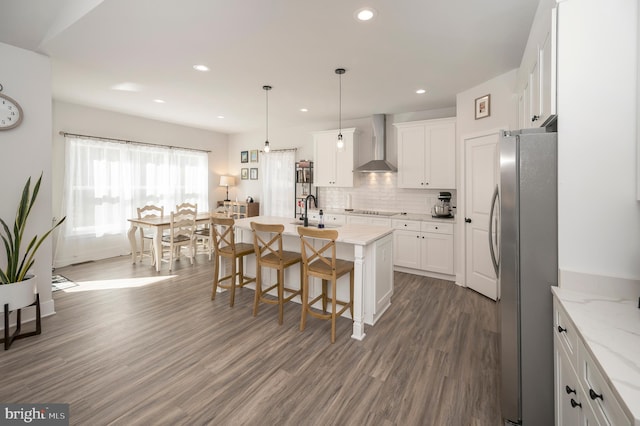 kitchen featuring white cabinets, stainless steel fridge, an island with sink, and wall chimney exhaust hood