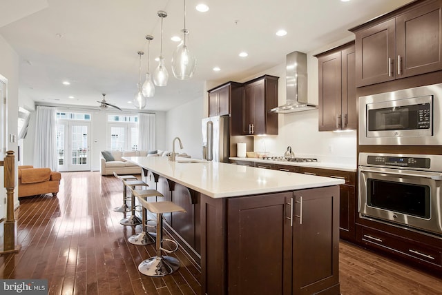 kitchen with stainless steel appliances, open floor plan, light countertops, wall chimney range hood, and an island with sink