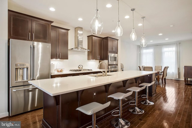 kitchen featuring a kitchen island with sink, stainless steel appliances, light countertops, wall chimney exhaust hood, and pendant lighting