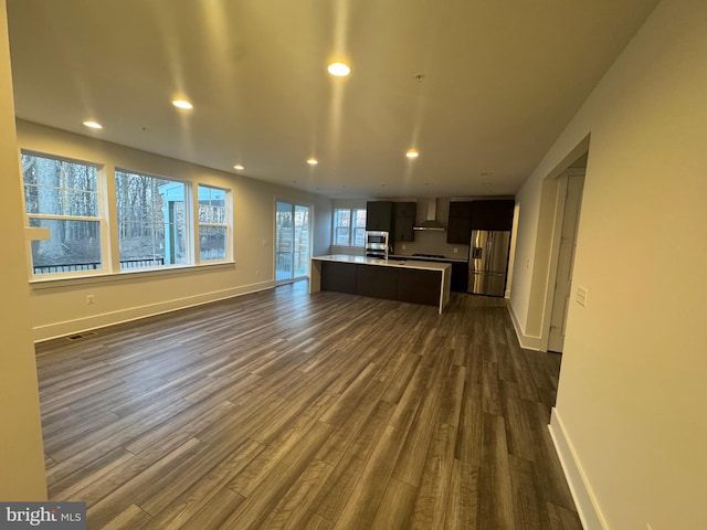 kitchen featuring open floor plan, light countertops, wall chimney range hood, stainless steel refrigerator with ice dispenser, and a center island with sink