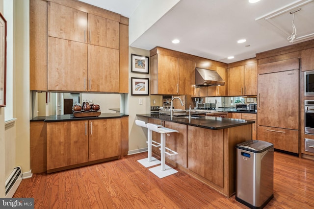 kitchen with built in appliances, light wood-style floors, baseboard heating, wall chimney exhaust hood, and dark countertops