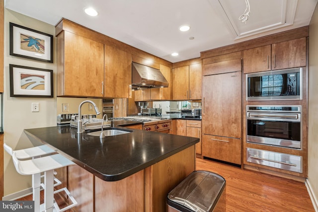 kitchen featuring a warming drawer, a sink, built in appliances, a peninsula, and wall chimney exhaust hood