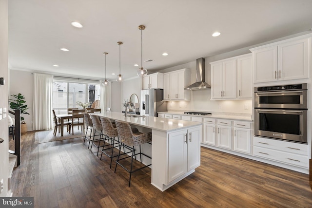 kitchen with light countertops, appliances with stainless steel finishes, white cabinets, an island with sink, and wall chimney exhaust hood