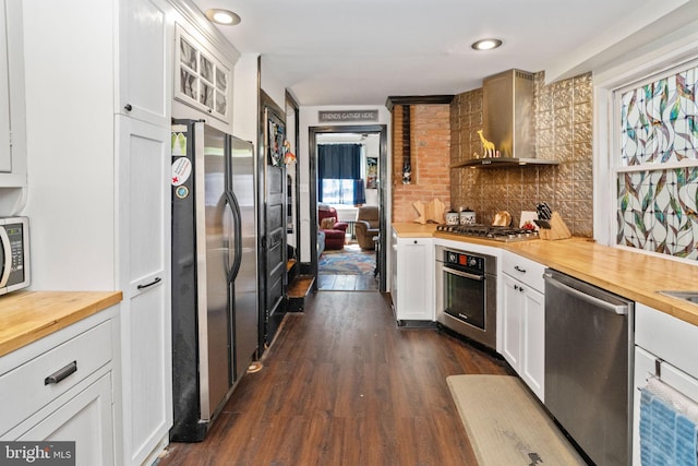 kitchen featuring appliances with stainless steel finishes, butcher block countertops, white cabinetry, dark wood-type flooring, and wall chimney range hood