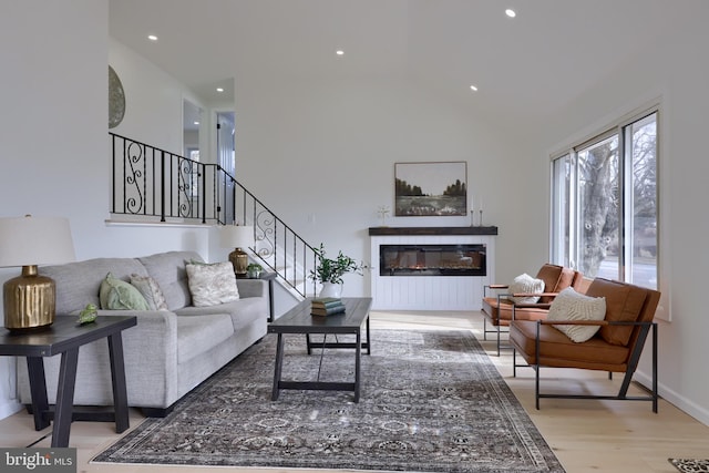 living area with light wood finished floors, recessed lighting, stairway, a high ceiling, and a glass covered fireplace