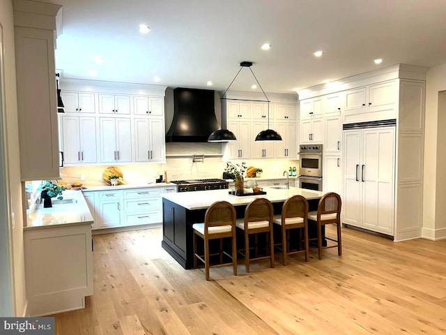 kitchen with light hardwood / wood-style flooring, a kitchen island with sink, a kitchen breakfast bar, wall chimney exhaust hood, and stainless steel double oven