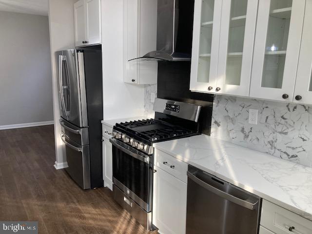 kitchen with wall chimney exhaust hood, appliances with stainless steel finishes, glass insert cabinets, and white cabinetry