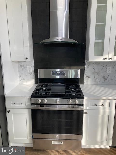 kitchen featuring white cabinets, wall chimney exhaust hood, glass insert cabinets, stainless steel gas range, and light countertops