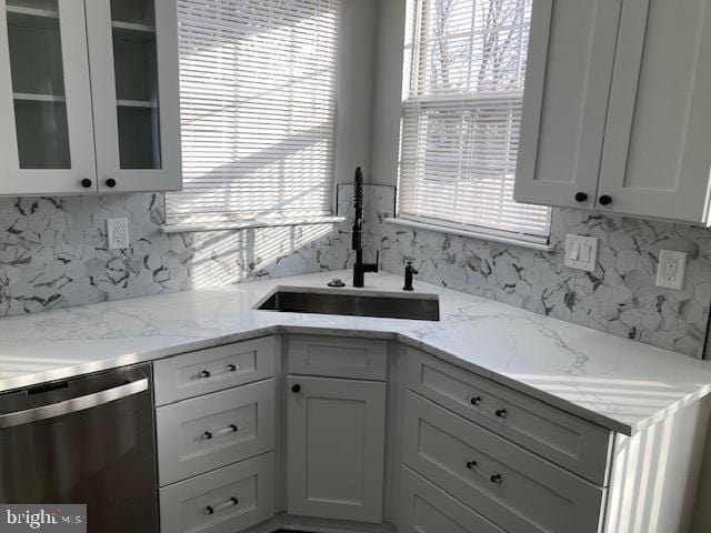 kitchen featuring stainless steel dishwasher, glass insert cabinets, white cabinetry, a sink, and light stone countertops