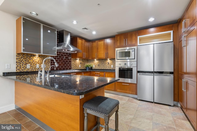 kitchen featuring dark stone countertops, wall chimney range hood, kitchen peninsula, and appliances with stainless steel finishes