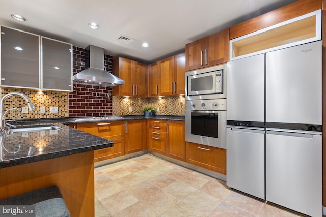 kitchen featuring sink, backsplash, dark stone counters, stainless steel appliances, and wall chimney exhaust hood