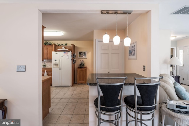 kitchen featuring visible vents, a kitchen breakfast bar, white fridge with ice dispenser, brown cabinetry, and dark countertops