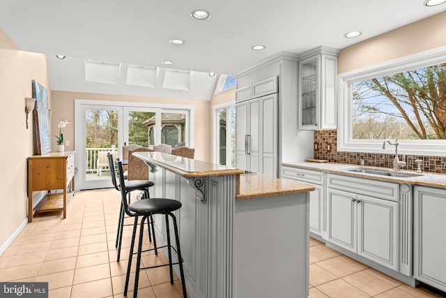 kitchen featuring light stone counters, a sink, backsplash, a center island, and glass insert cabinets