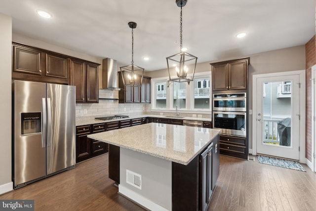 kitchen with wall chimney exhaust hood, light stone counters, hanging light fixtures, a kitchen island, and stainless steel appliances