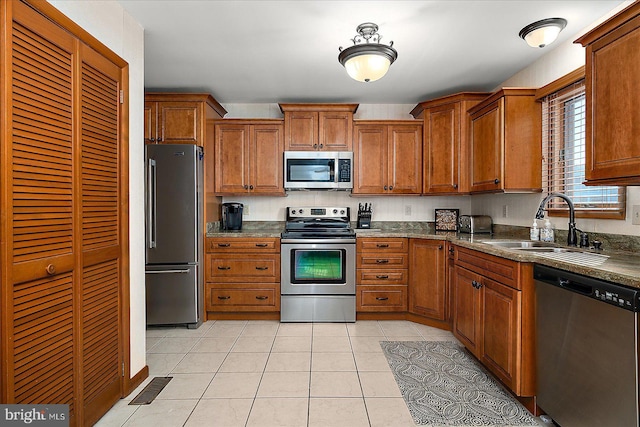 kitchen featuring dark countertops, appliances with stainless steel finishes, brown cabinets, a sink, and light tile patterned flooring