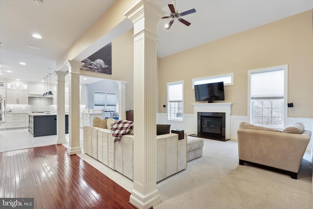 living room featuring a wainscoted wall, ceiling fan, a premium fireplace, light wood-style flooring, and ornate columns