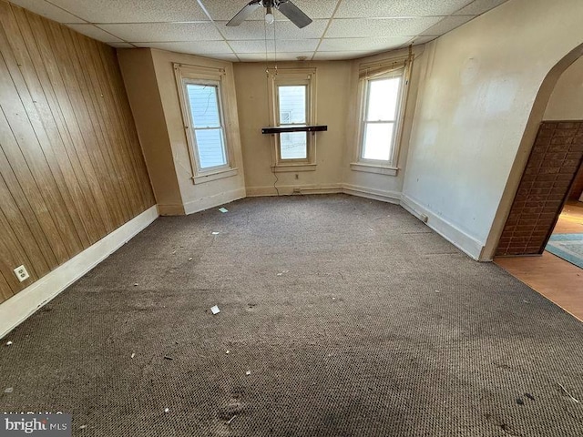 empty room with ceiling fan, wood walls, carpet floors, and a drop ceiling