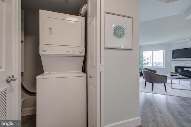 clothes washing area with laundry area, baseboards, a glass covered fireplace, wood finished floors, and stacked washing maching and dryer