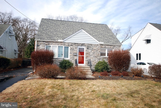 cape cod house featuring a shingled roof, a front yard, stone siding, and fence