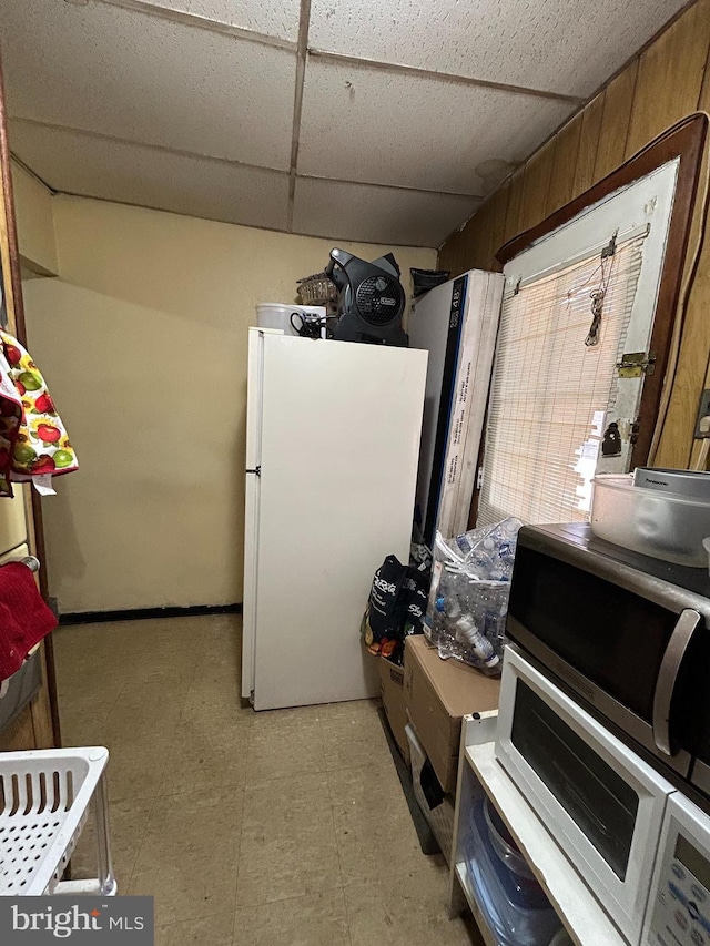 kitchen featuring a drop ceiling, light floors, wood walls, and freestanding refrigerator