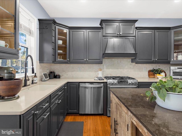 kitchen with light wood finished floors, glass insert cabinets, custom range hood, stainless steel appliances, and a sink