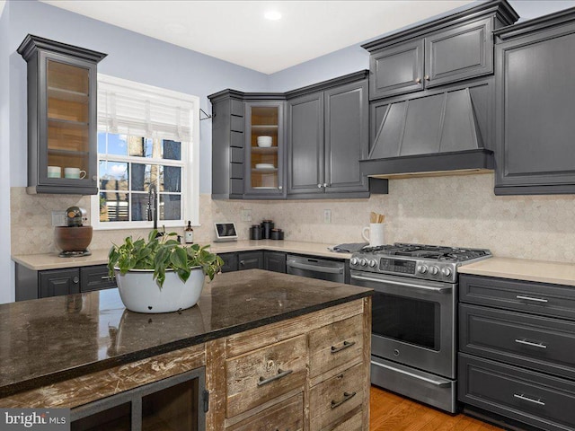 kitchen with backsplash, glass insert cabinets, premium range hood, dark stone countertops, and appliances with stainless steel finishes