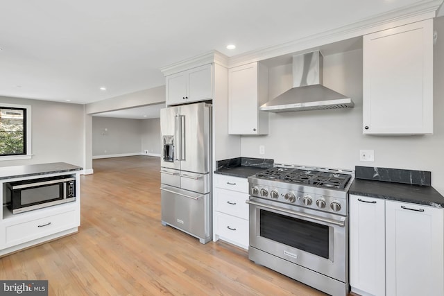 kitchen with high quality appliances, dark stone countertops, white cabinets, wall chimney exhaust hood, and light hardwood / wood-style flooring