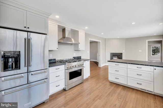 kitchen with wall chimney range hood, dark stone countertops, high end appliances, white cabinets, and light wood-type flooring