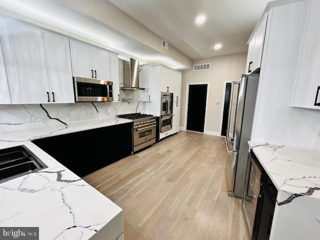 kitchen featuring stainless steel appliances, wall chimney exhaust hood, light stone countertops, and white cabinets