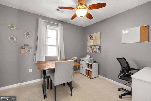 home office with a textured ceiling, light carpet, visible vents, a ceiling fan, and baseboards