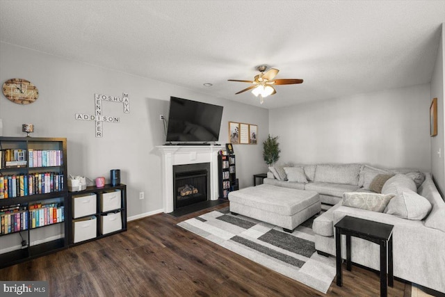 living area featuring a textured ceiling, a fireplace with flush hearth, a ceiling fan, baseboards, and dark wood-style floors