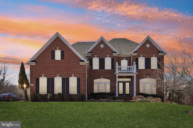 colonial-style house featuring a balcony, roof with shingles, a front yard, and brick siding