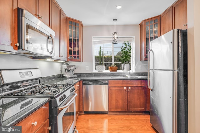 kitchen with glass insert cabinets, appliances with stainless steel finishes, brown cabinets, light wood-style floors, and pendant lighting