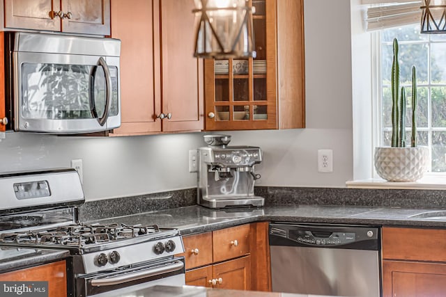 kitchen featuring appliances with stainless steel finishes, brown cabinets, glass insert cabinets, and dark stone countertops