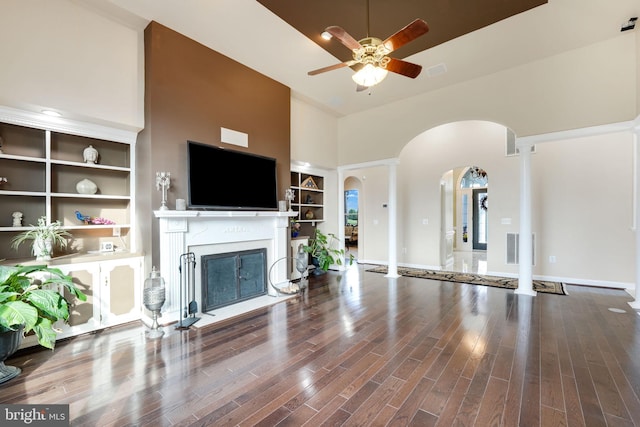 unfurnished living room featuring arched walkways, ceiling fan, wood finished floors, a glass covered fireplace, and ornate columns