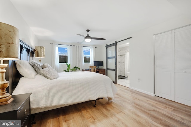 bedroom featuring a barn door, light wood-style floors, ceiling fan, ensuite bath, and baseboards