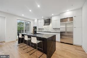 kitchen featuring stainless steel built in refrigerator, a breakfast bar, light hardwood / wood-style flooring, a center island with sink, and white cabinets