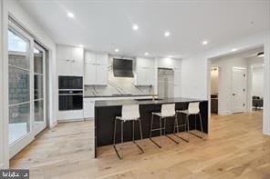 kitchen with a breakfast bar, ventilation hood, white cabinetry, a kitchen island with sink, and light hardwood / wood-style flooring