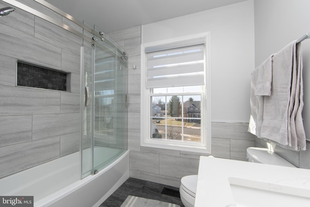 bathroom featuring tile walls, shower / bath combination with glass door, and toilet