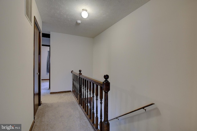 hallway with a textured ceiling, light colored carpet, visible vents, baseboards, and an upstairs landing