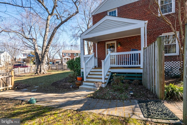 doorway to property featuring a porch, brick siding, and fence