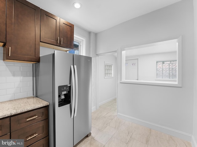 kitchen featuring dark brown cabinetry, light stone counters, tasteful backsplash, a wealth of natural light, and stainless steel fridge with ice dispenser